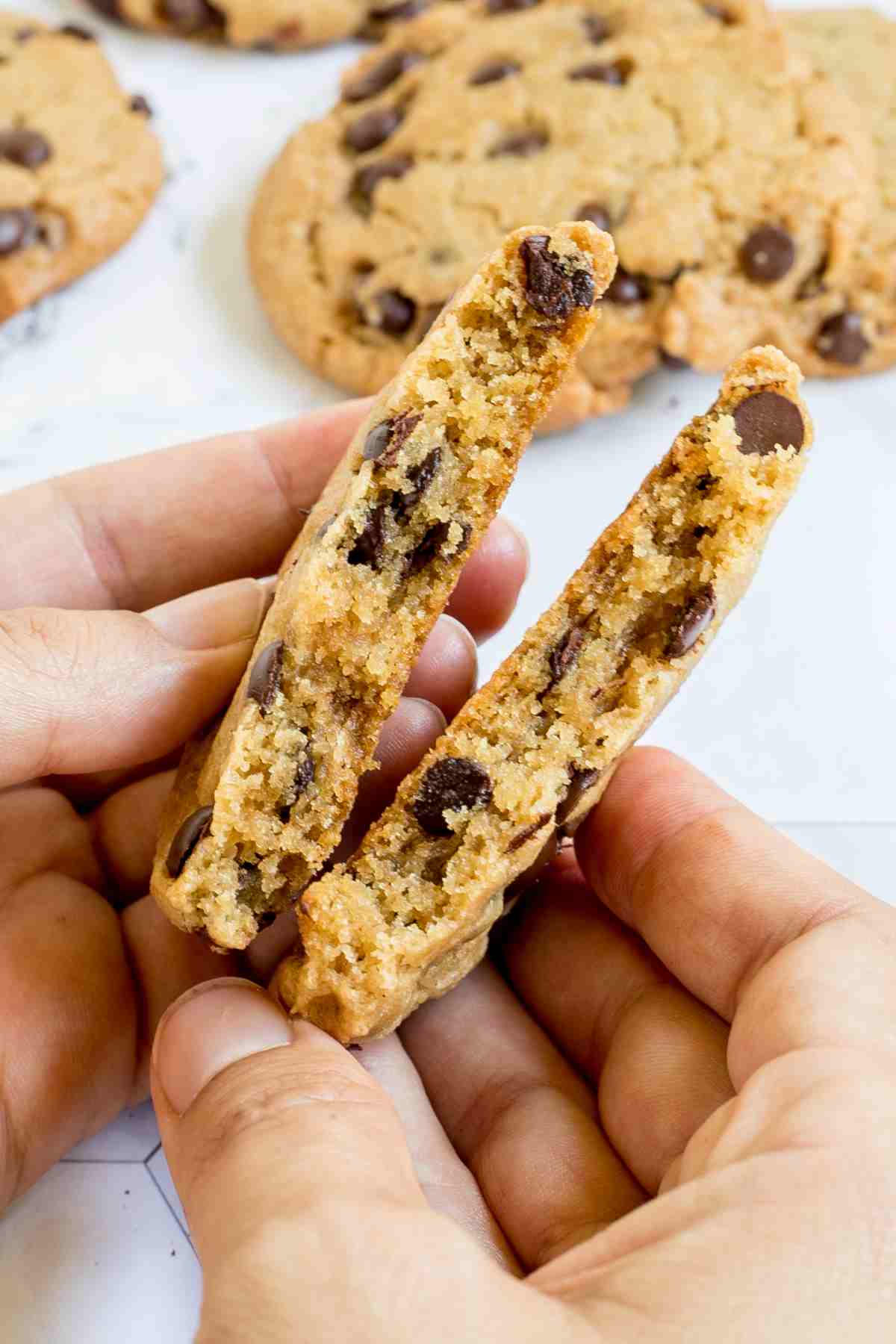 A hand is breaking a chocolate chip cookie in the middle. More cookies are in the background.