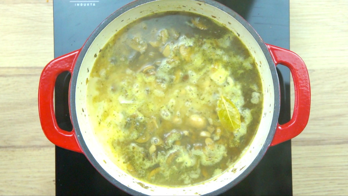 Red-white enameled Dutch oven with a thin brown soup full of mushrooms and two bay leaves swimming on top. There is a creamy bit on the side indicating something white just had been added to the soup.