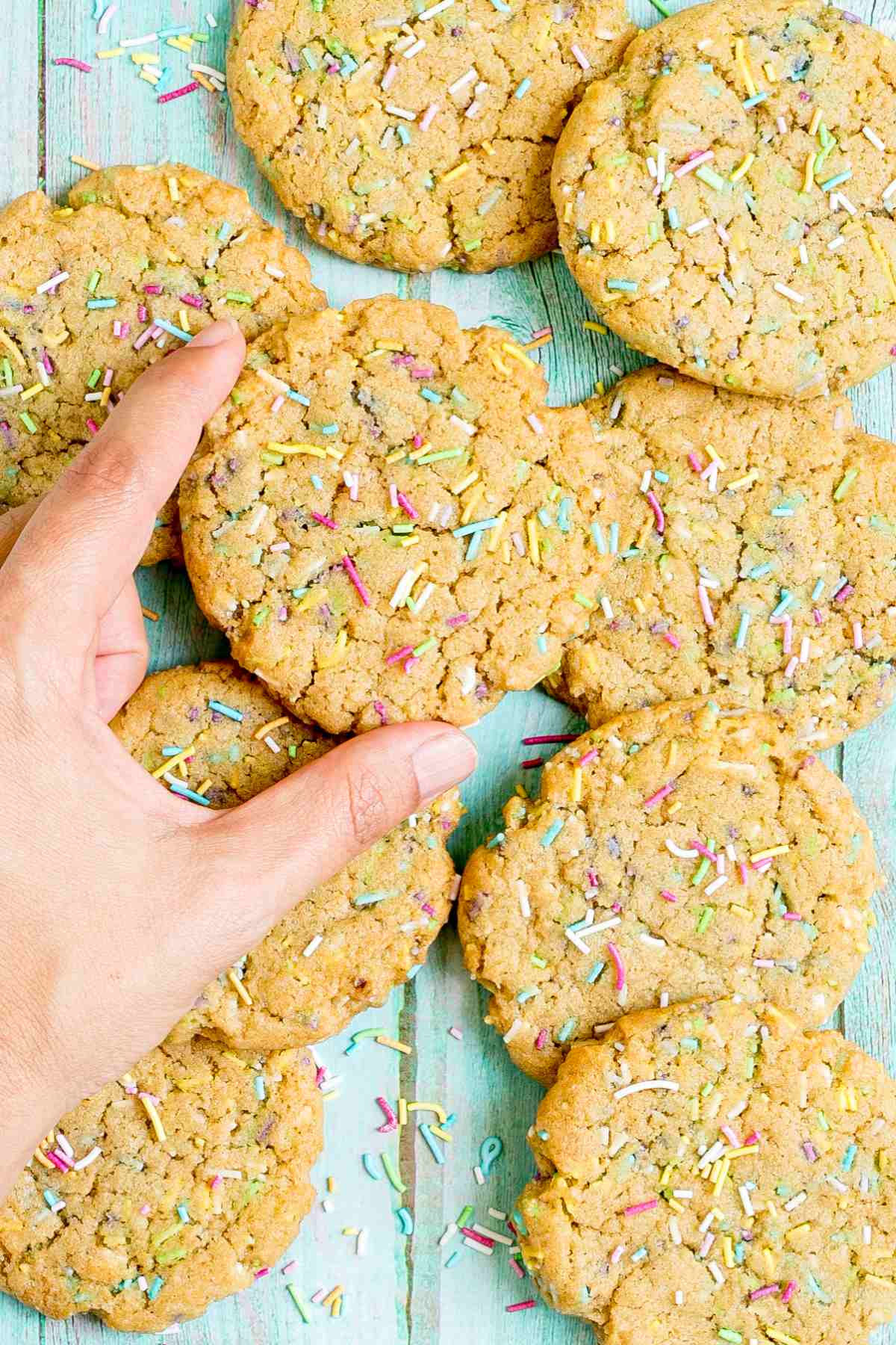 Light brown cookies with rainbow sprinkles on a blue wooden surface. A hand is taking one from the top.