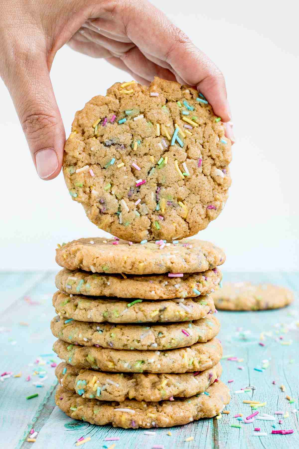 A stack of light brown cookies with rainbow sprinkles on a blue wooden surface. A hand is taking one from the top.