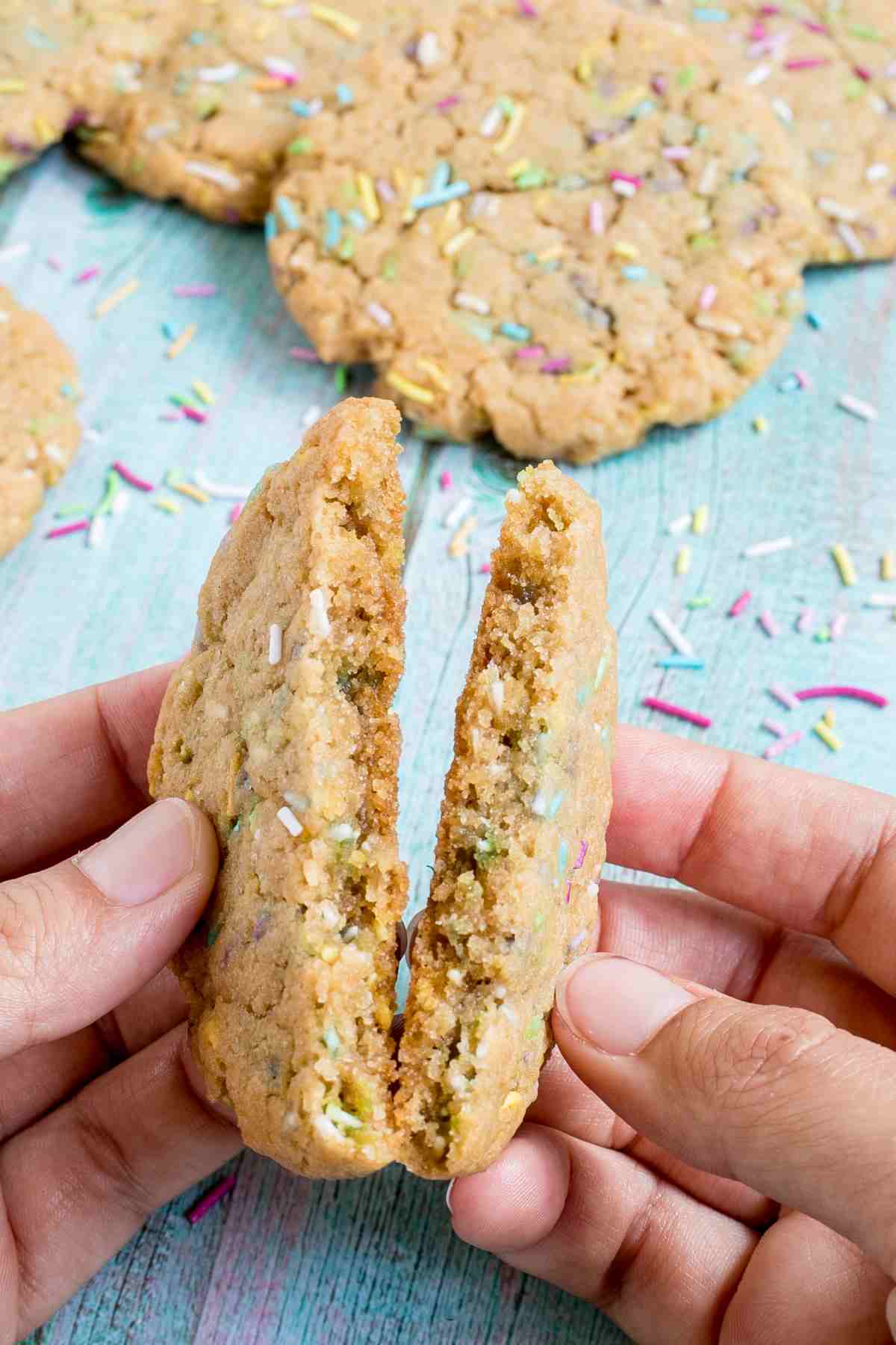 A hand is breaking a light brown cookie with rainbow sprinkles in half to show the inner texture. More cookies are behind it.