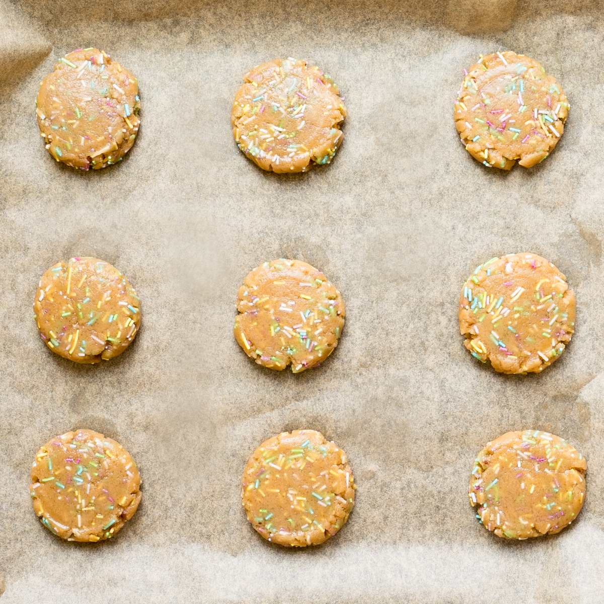 Uncooked light brown cookies with rainbow sprinkles on parchment paper.