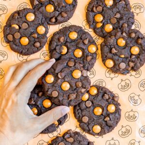 Dark brown cookies with orange m&ms, and chocolate chips on an orange surface with a pumpkin pattern. A hand is grabbing one.