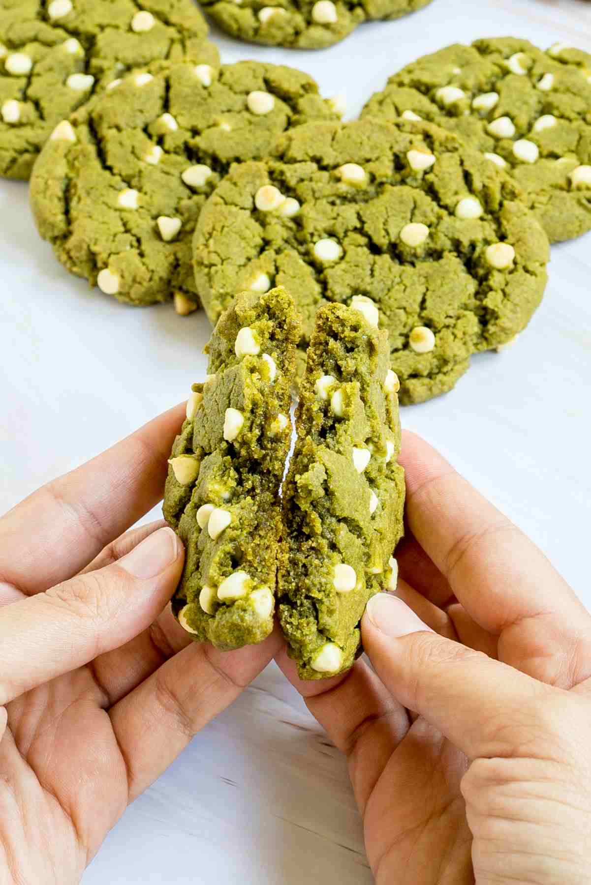 A hand is breaking a green cookie with white chocolate chips in half showing the texture. More cookies are in the background.