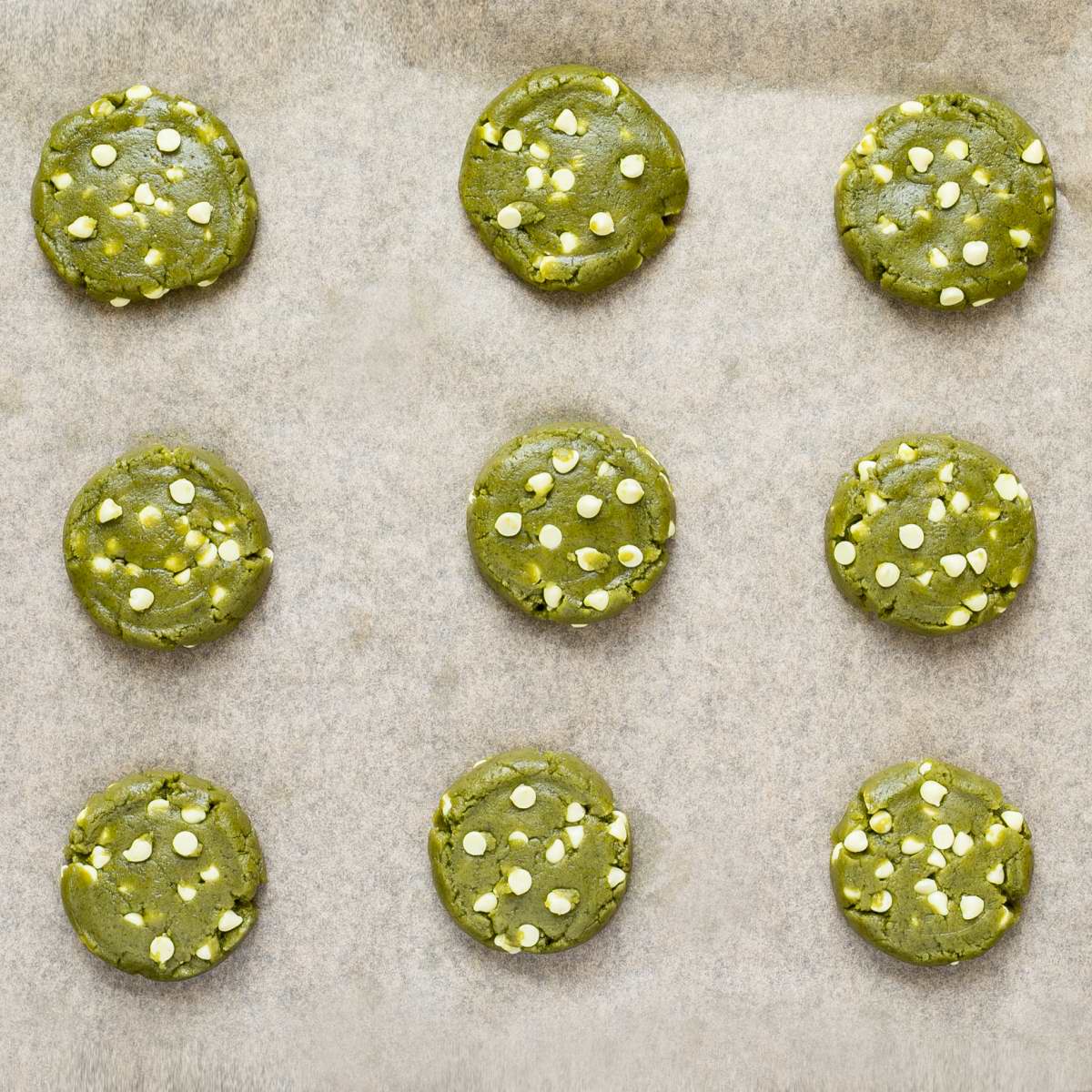 Unbaked round vibrant green cookie batters with white chocolate chips on parchment paper neatly placed away from each other.