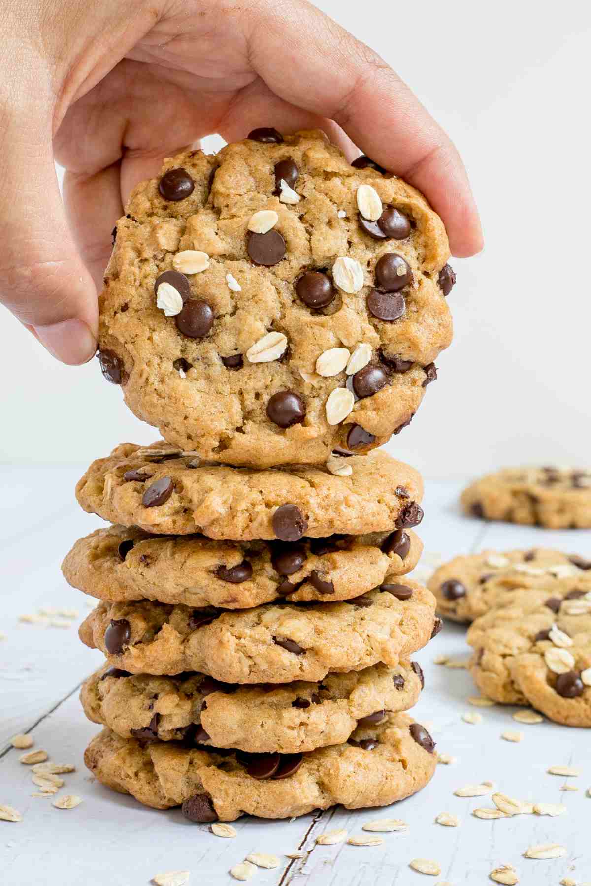 A stack of cookies with oats and chocolate chips on a wooden surface. More oats are sprinkled around them. A hand is taking the top one.