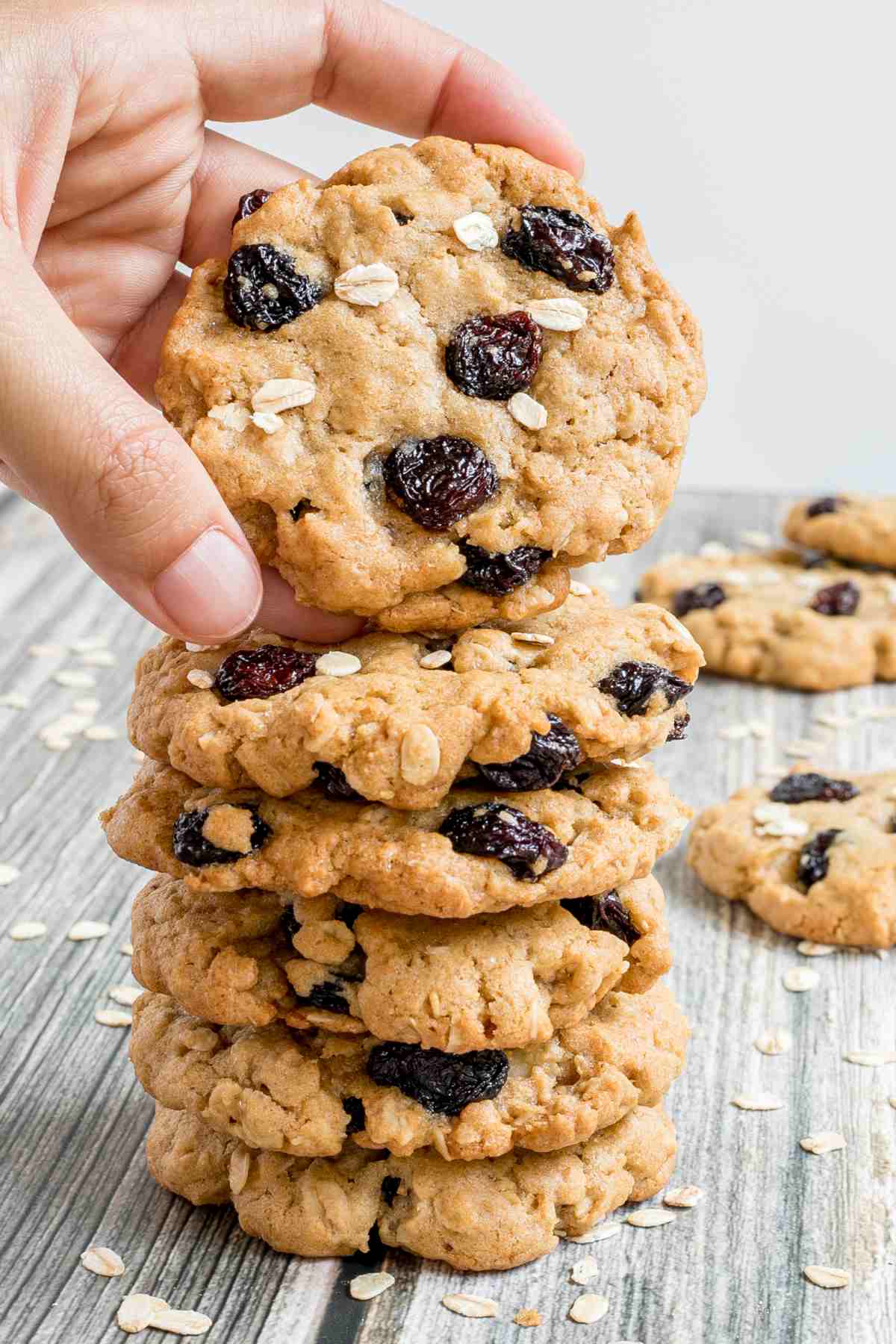 A stack of cookies with oats and raisins on a wooden surface. More oats are sprinkled around them. A hand is taking one from the top.