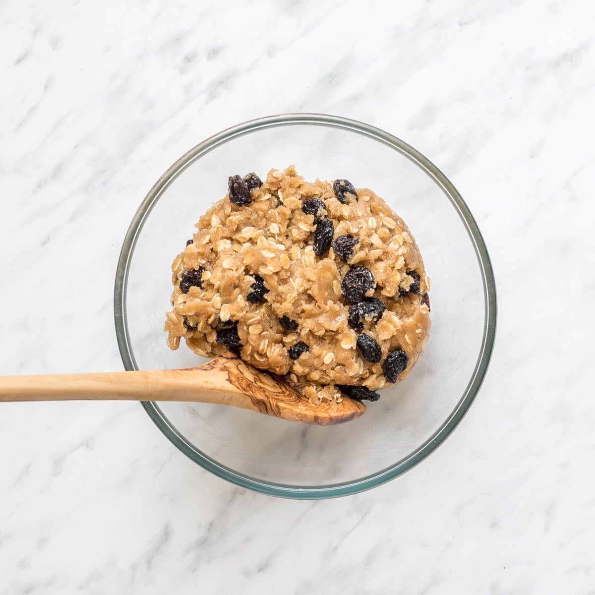A glass bowl with a light brown cookie batter with oats and raisins.