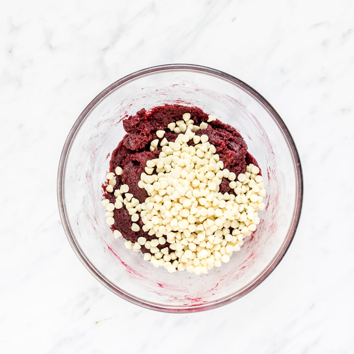 A glass bowl with red cookie dough and a heap of white chocolate chips.