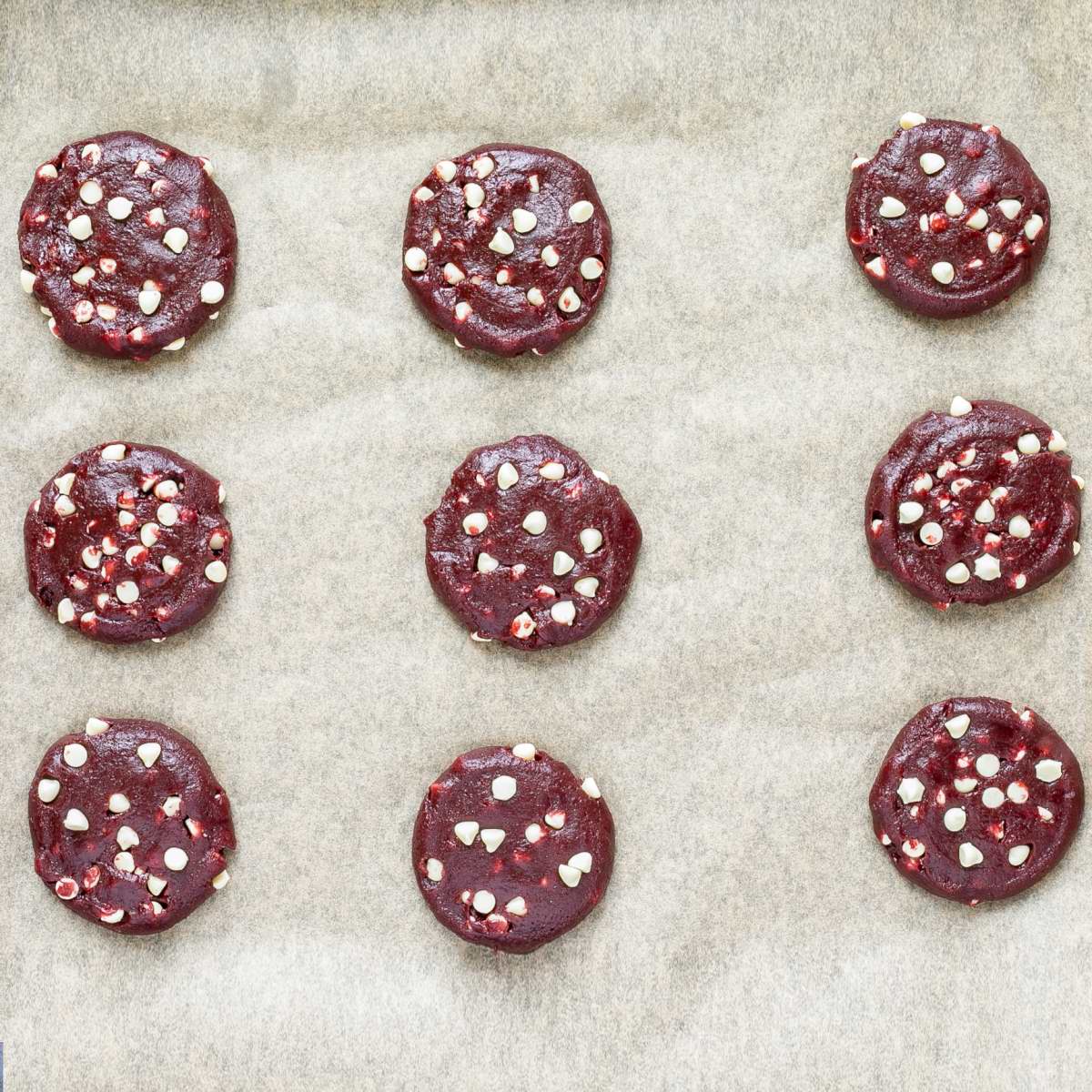 Unbaked round vibrant red cookie batters with white chocolate chips on parchment paper neatly placed away from each other.