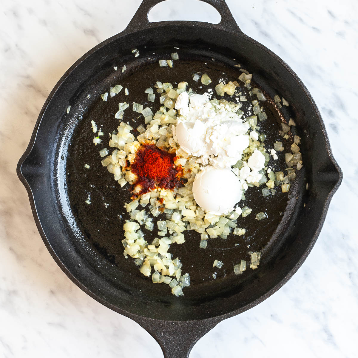 A black cast iron skillet from above with minced onion, heaps of white powder and a small heap of red paprika powder