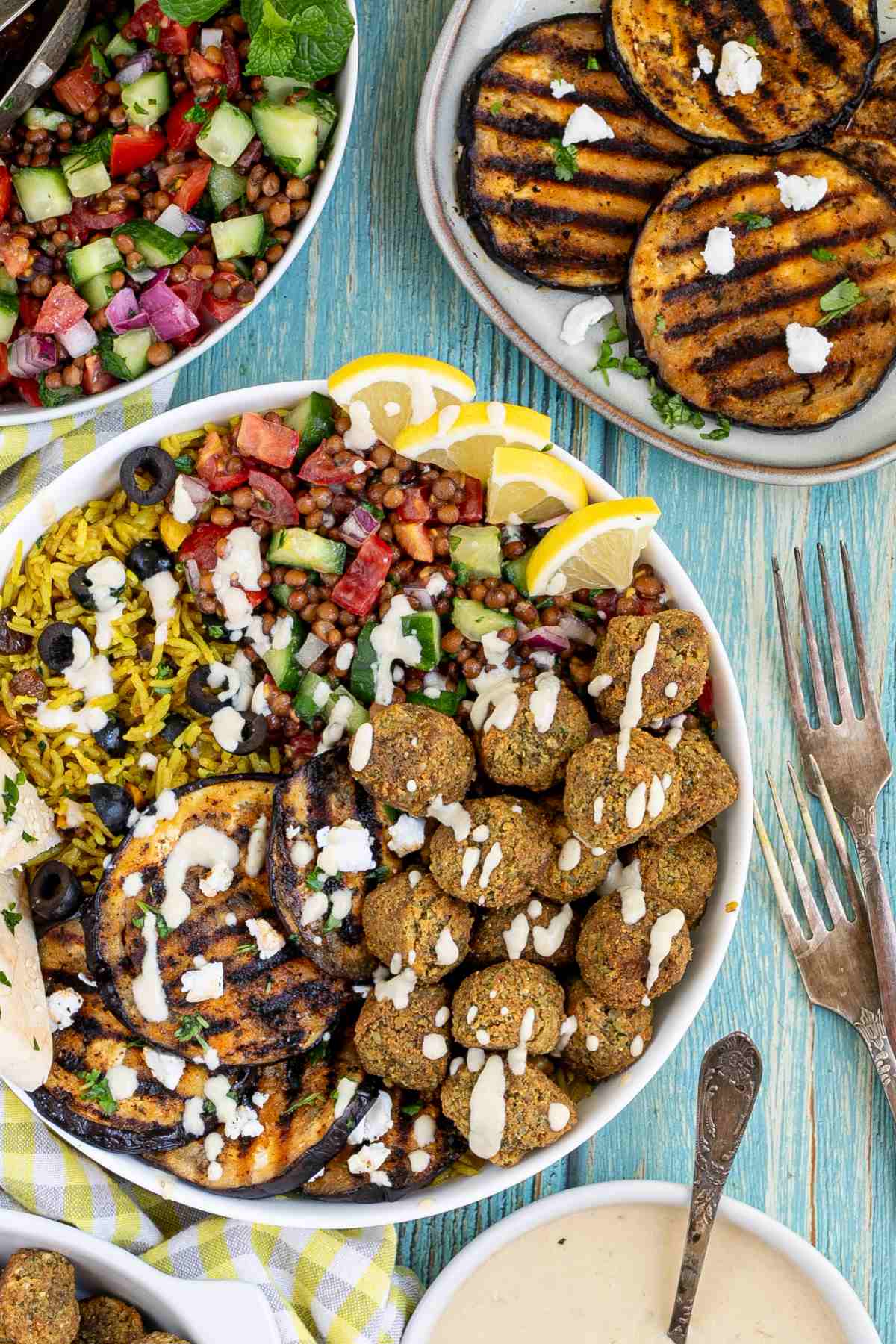 A large white bowl with falafel balls, grilled eggplant, yellow rice and chopped tomato and cucumber salad with lentils drizzled with a white sauce. The remaining ingredients are in bowls around the main one.