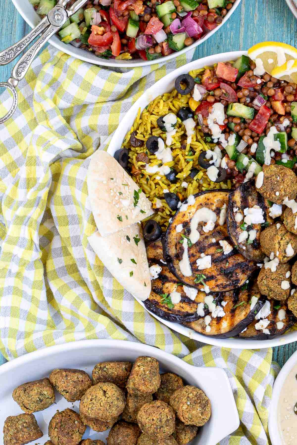 A large white bowl with falafel balls, grilled eggplant, yellow rice and chopped tomato and cucumber salad with lentils drizzled with a white sauce. The remaining ingredients are in bowls around the main one.