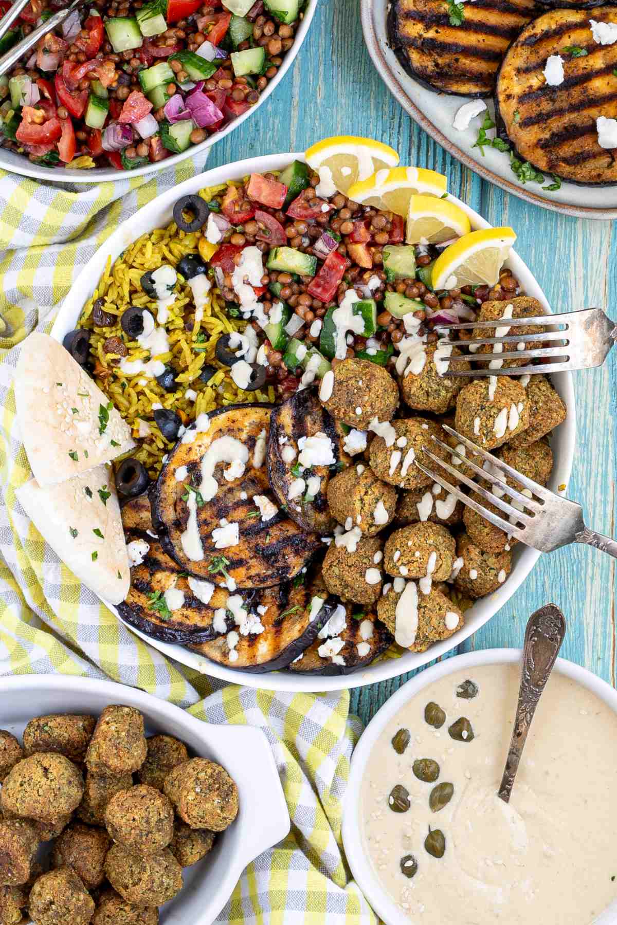 A large white bowl with falafel balls, grilled eggplant, yellow rice and chopped tomato and cucumber salad with lentils drizzled with a white sauce. 