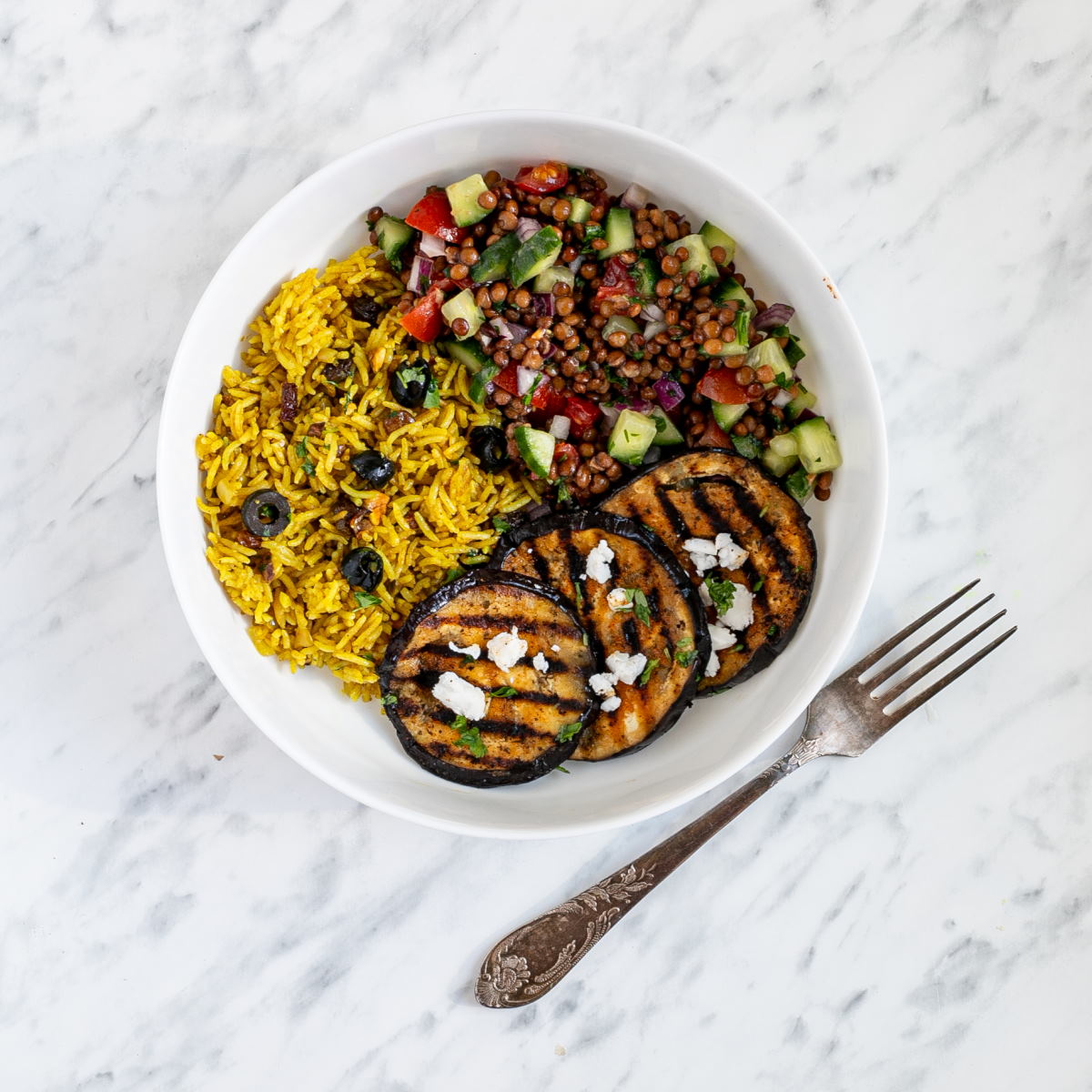 A large white bowl with grilled eggplant, yellow rice and chopped tomato and cucumber salad with lentils.