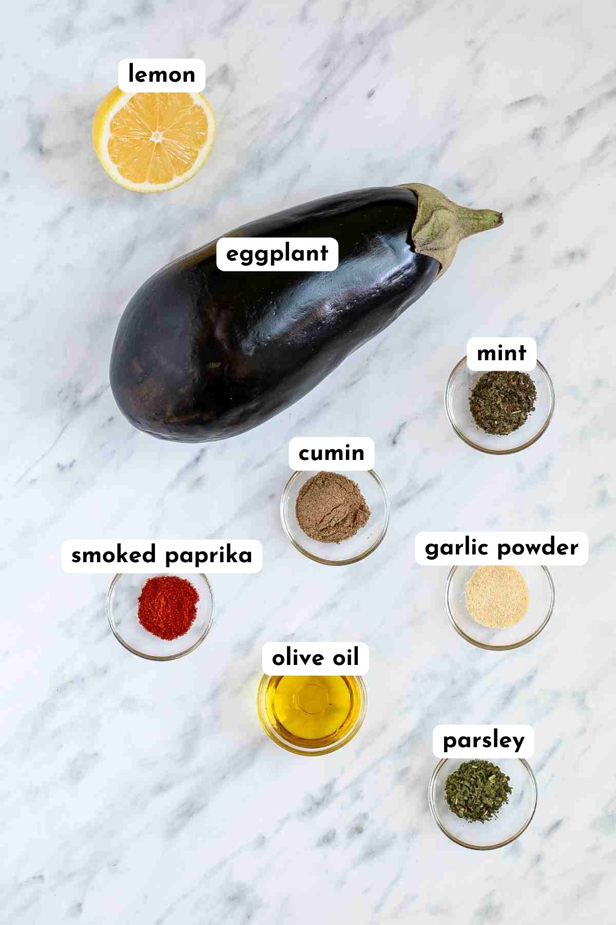 Ingredients of grilled eggplant in small glass bowls next to a large eggplant like mint, cumin, paprika powder, oil, garlic powder and parsley.