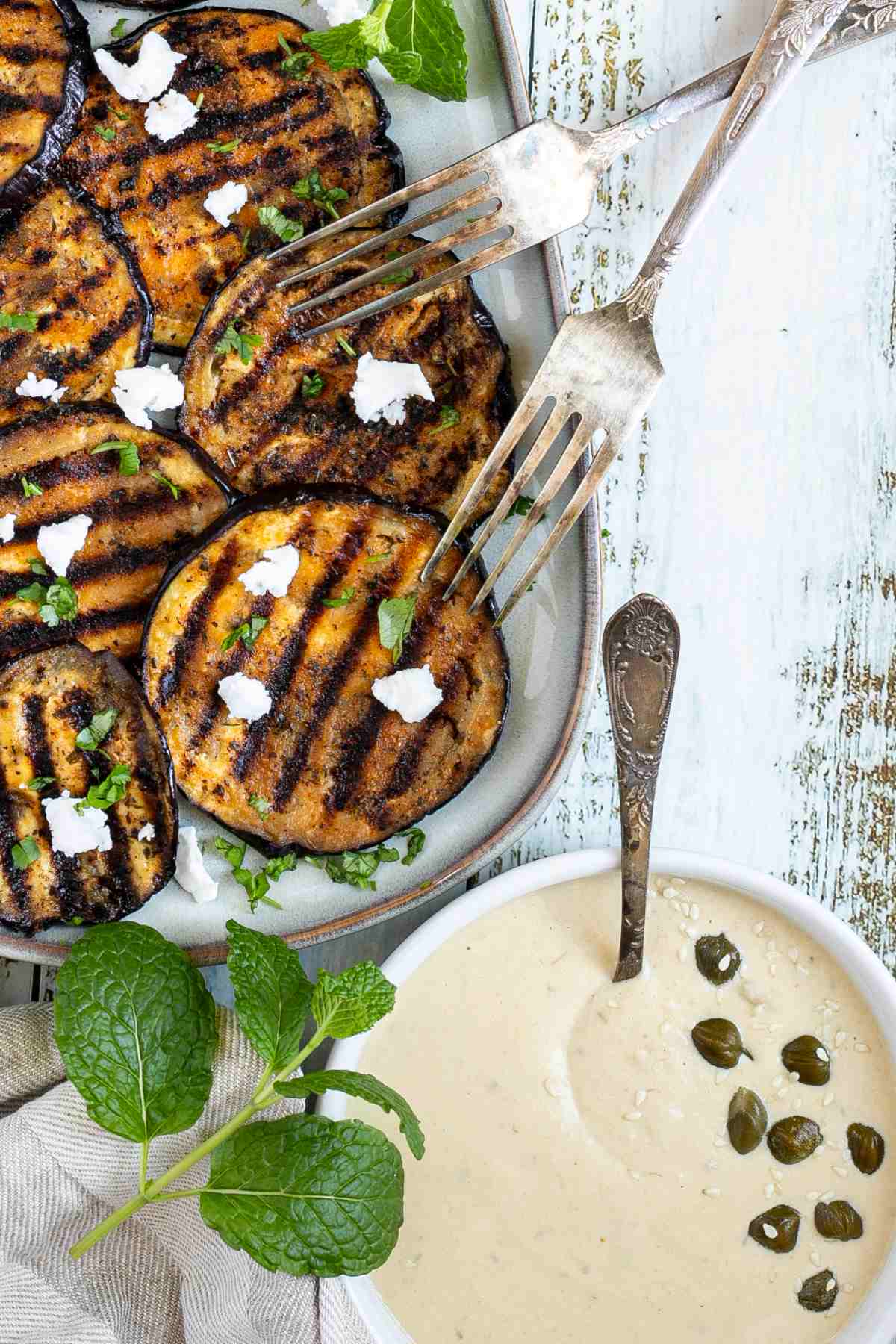 Large grilled slices of eggplant sprinkled with fresh herbs and crumbled cheese served on a light blue plate. A light brown sauce is placed next to it.