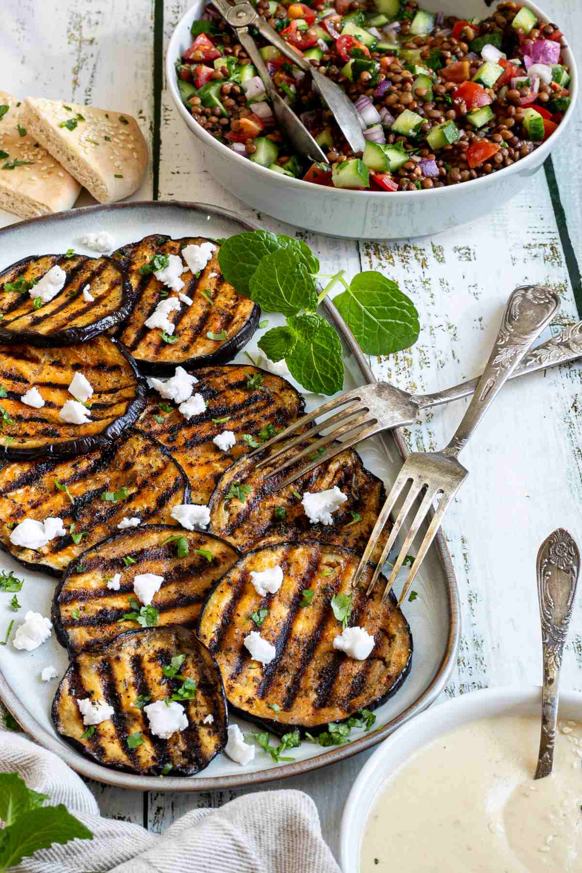 Large grilled slices of eggplant sprinkled with fresh herbs and crumbled cheese served on a light blue plate. A chopped mixed salad and a light brown sauce are next to it.