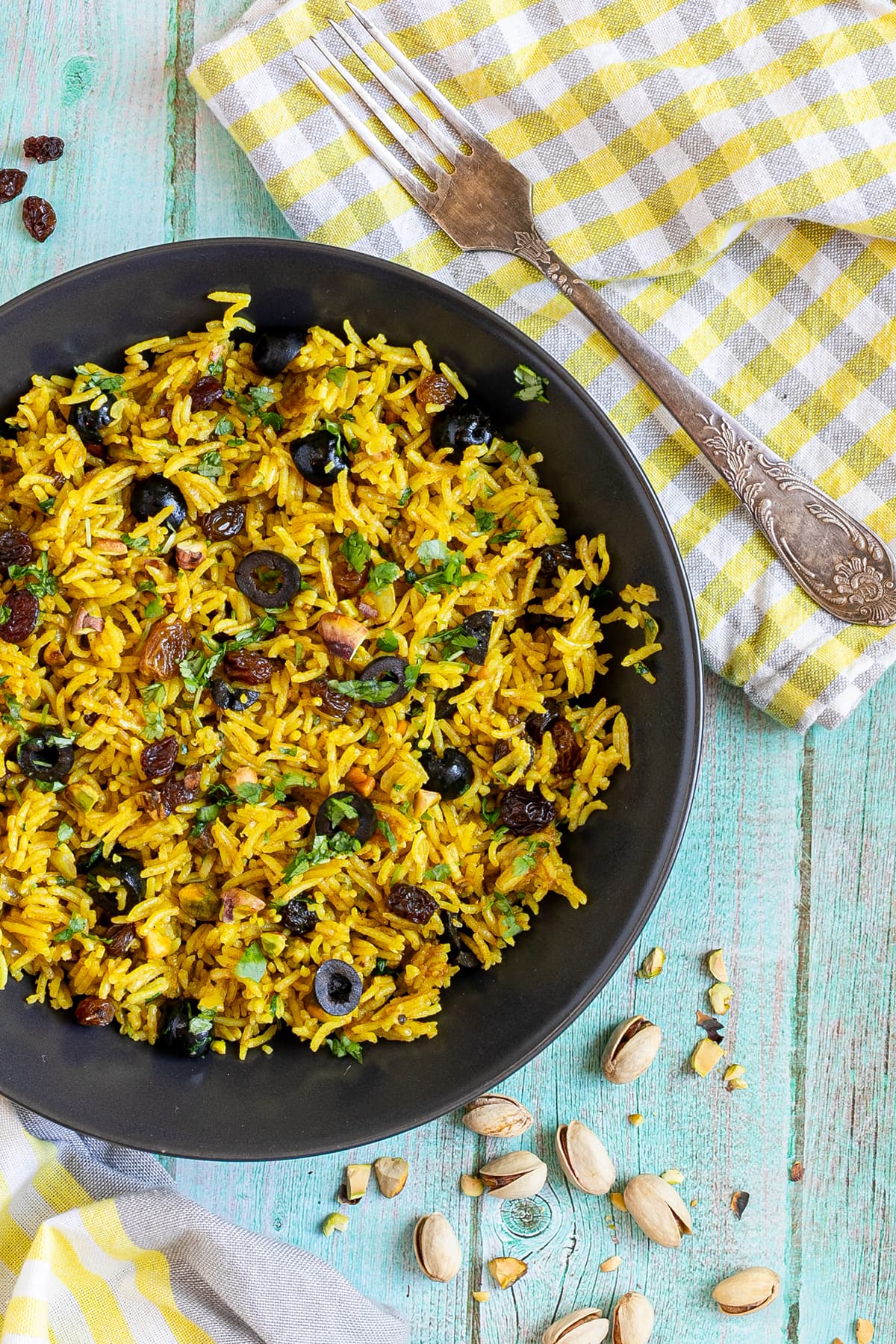 Yellow rice with raisins and pistachios and green herbs in a black bowl. More dry ingredients are scattered around it.