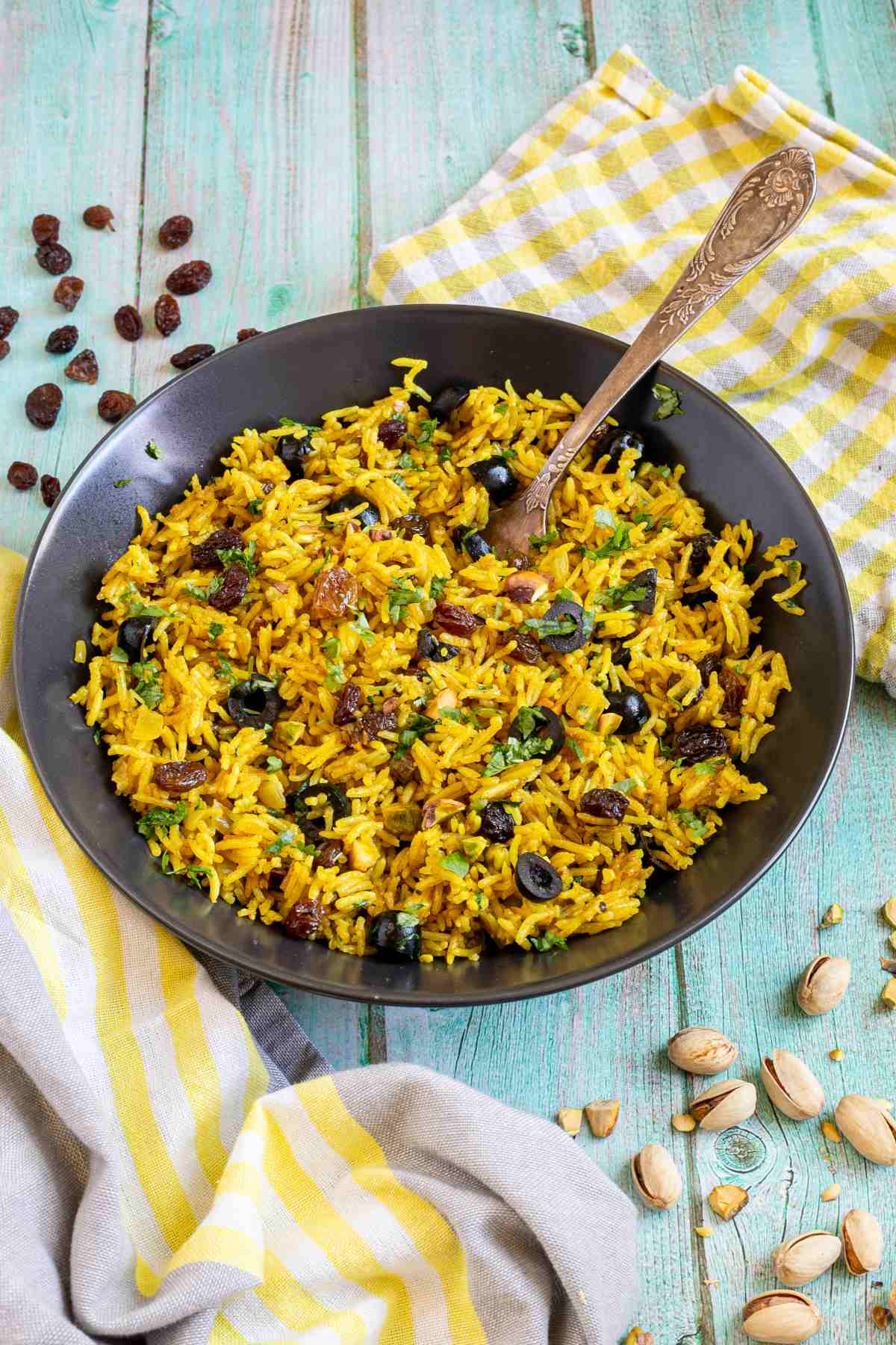 Yellow rice with raisins and pistachios and green herbs in a black bowl. More dry ingredients are scattered around it.