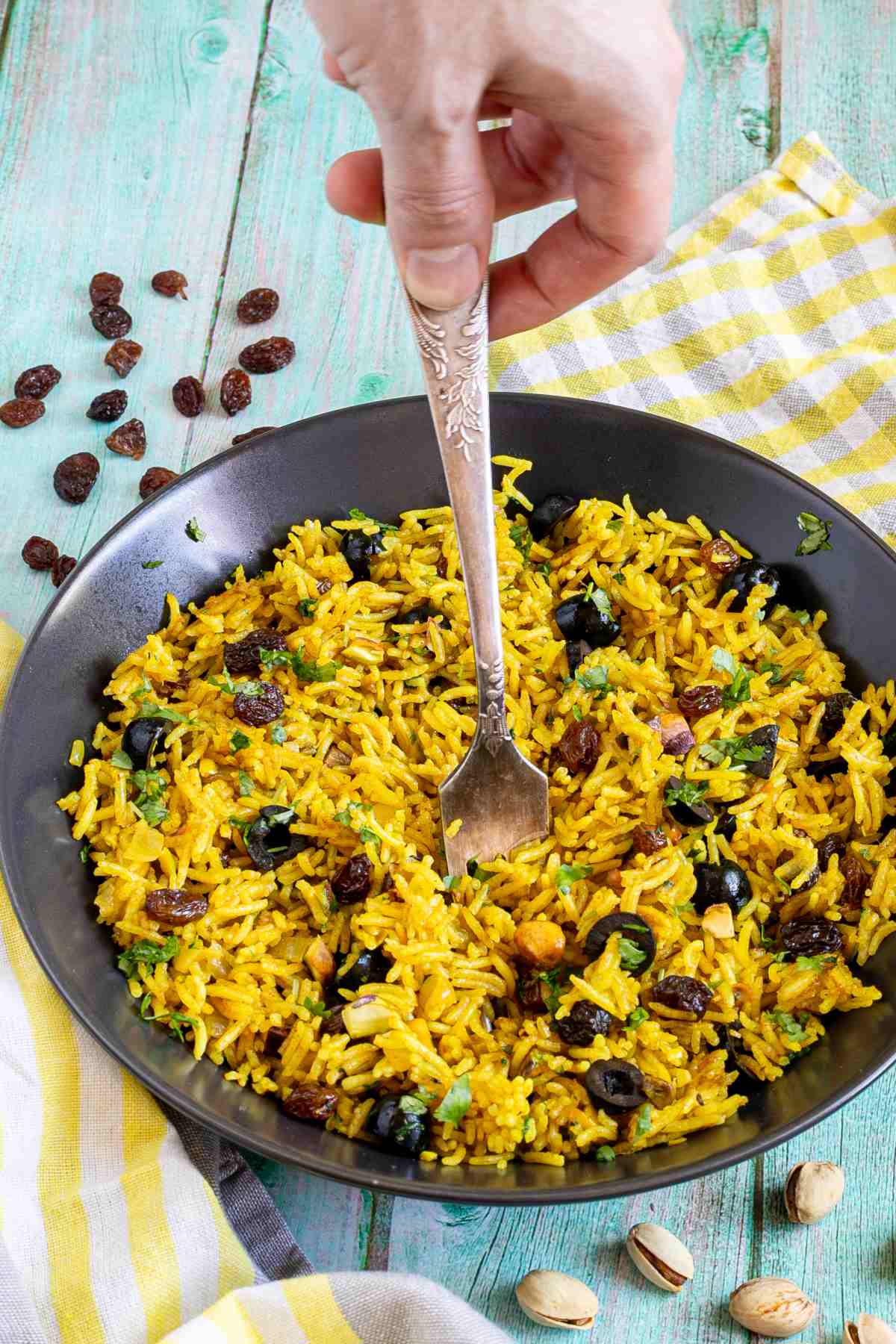 Yellow rice with raisins and pistachios and green herbs in a black bowl. A hand is holding a spoon to take some.