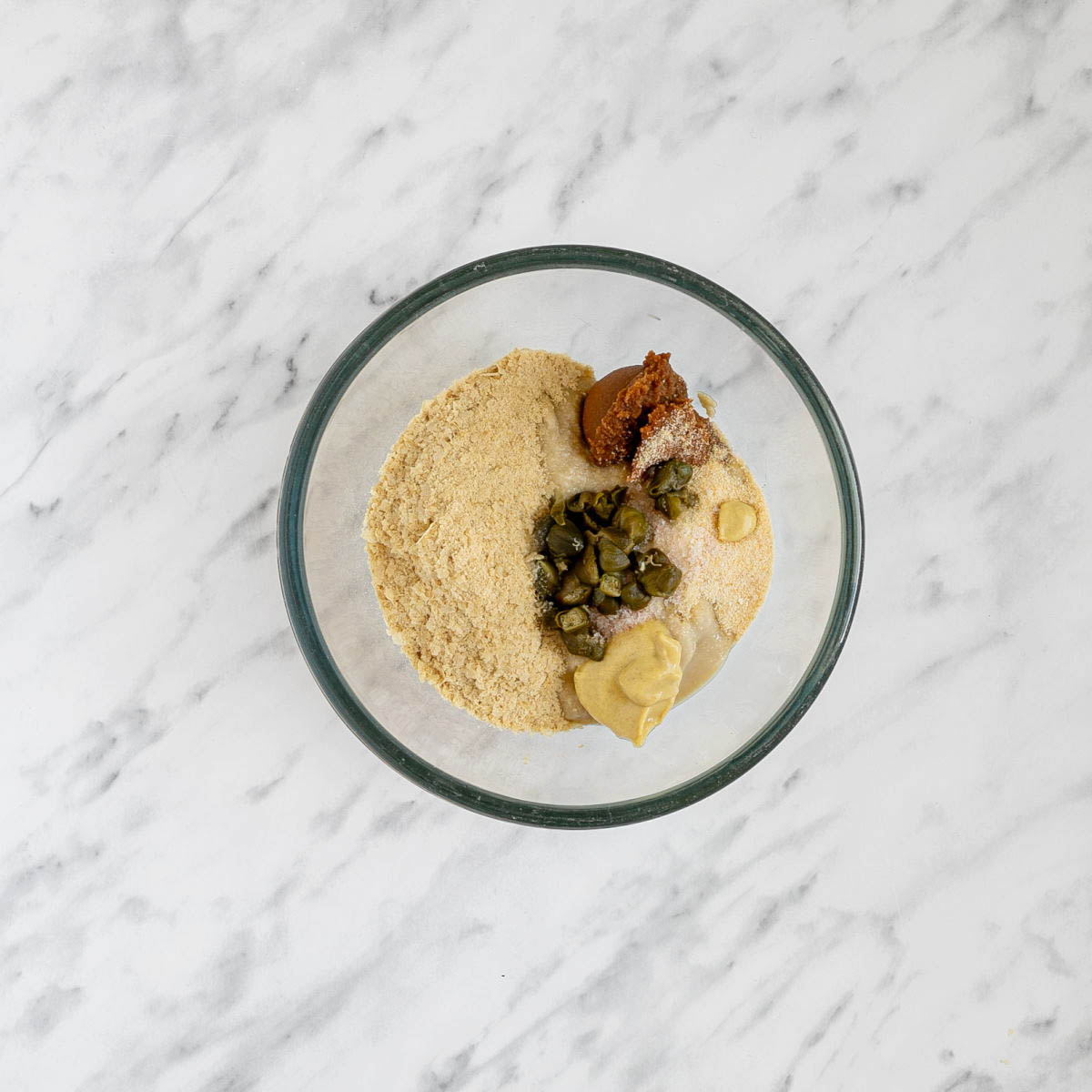 Glass bowl with light brown thick sauce, capers, dark brown paste, salt, and other ingredients.