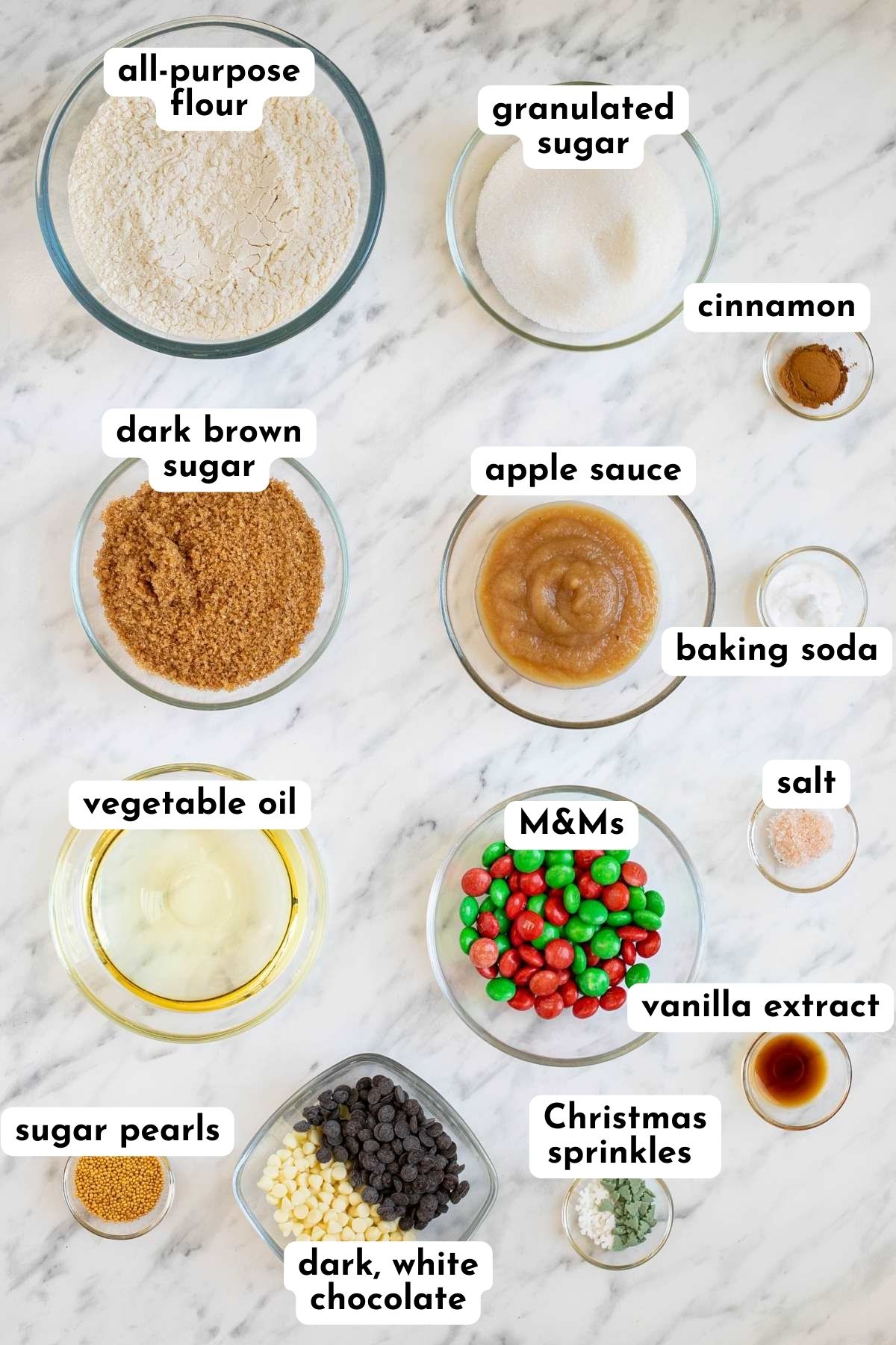 Ingredients of Christmas cookies in small glass bowls like flour, brown sugar, white sugar, apple sauce, oil, salt, cinnamon, vanilla extract, baking soda, green and red candies, white and green sprinkles, and white and dark chocolate chips.