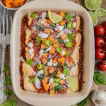 A casserole pan with rolled tortillas covered in red sauce and topped with diced roasted butternut squash, chili pepper slices, cilantro leaves, lime slices, sour cream and grated cheese.
