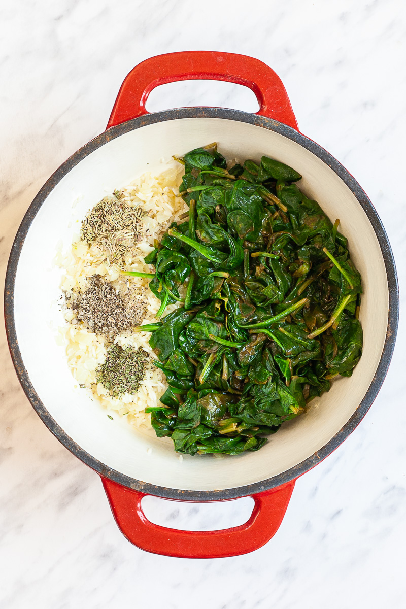 Wilted spinach leaves, and small heaps of dried herbs on sauteed onions in a red white enameled Dutch oven.