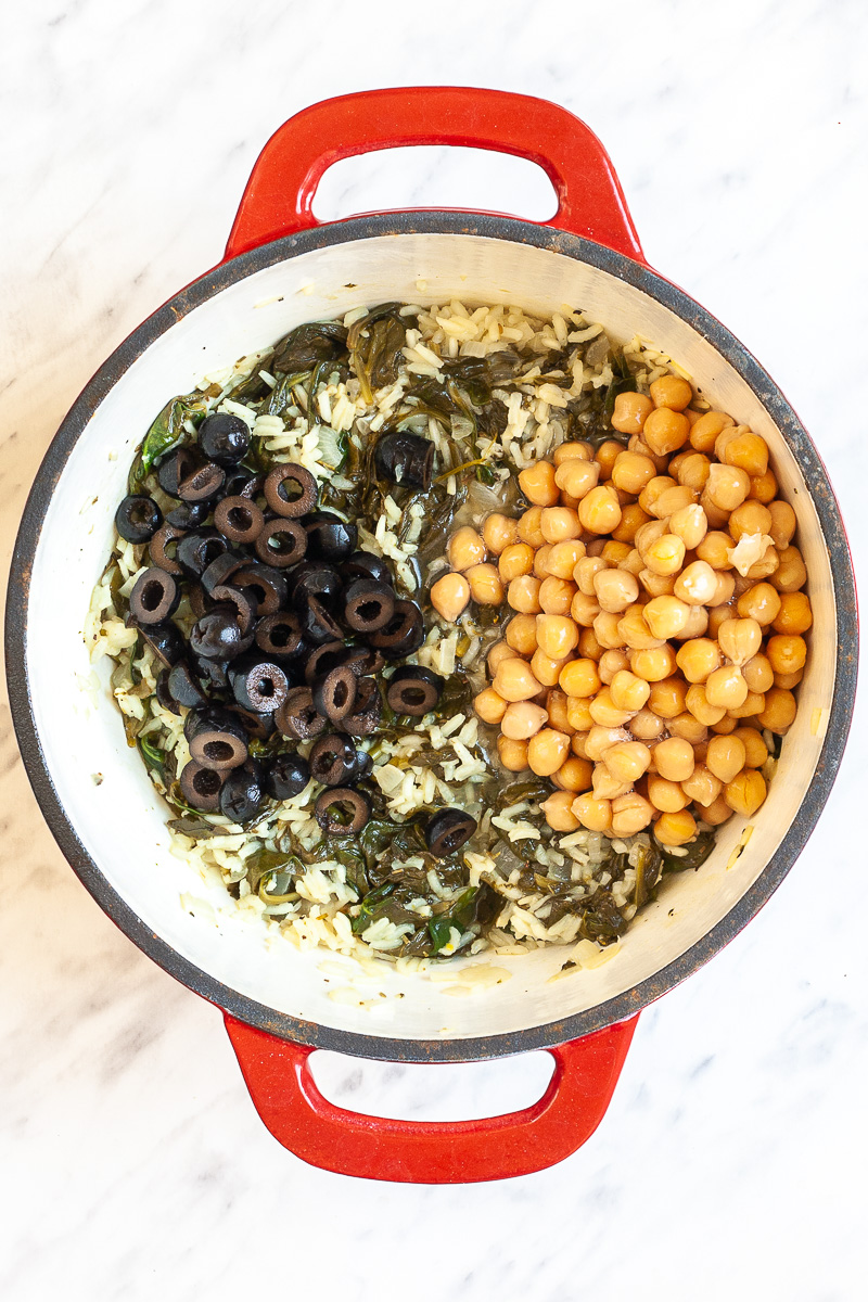 Rice, wilted spinach in an enameled red white Dutch oven topped with heaps of black olive slices and chickpeas.