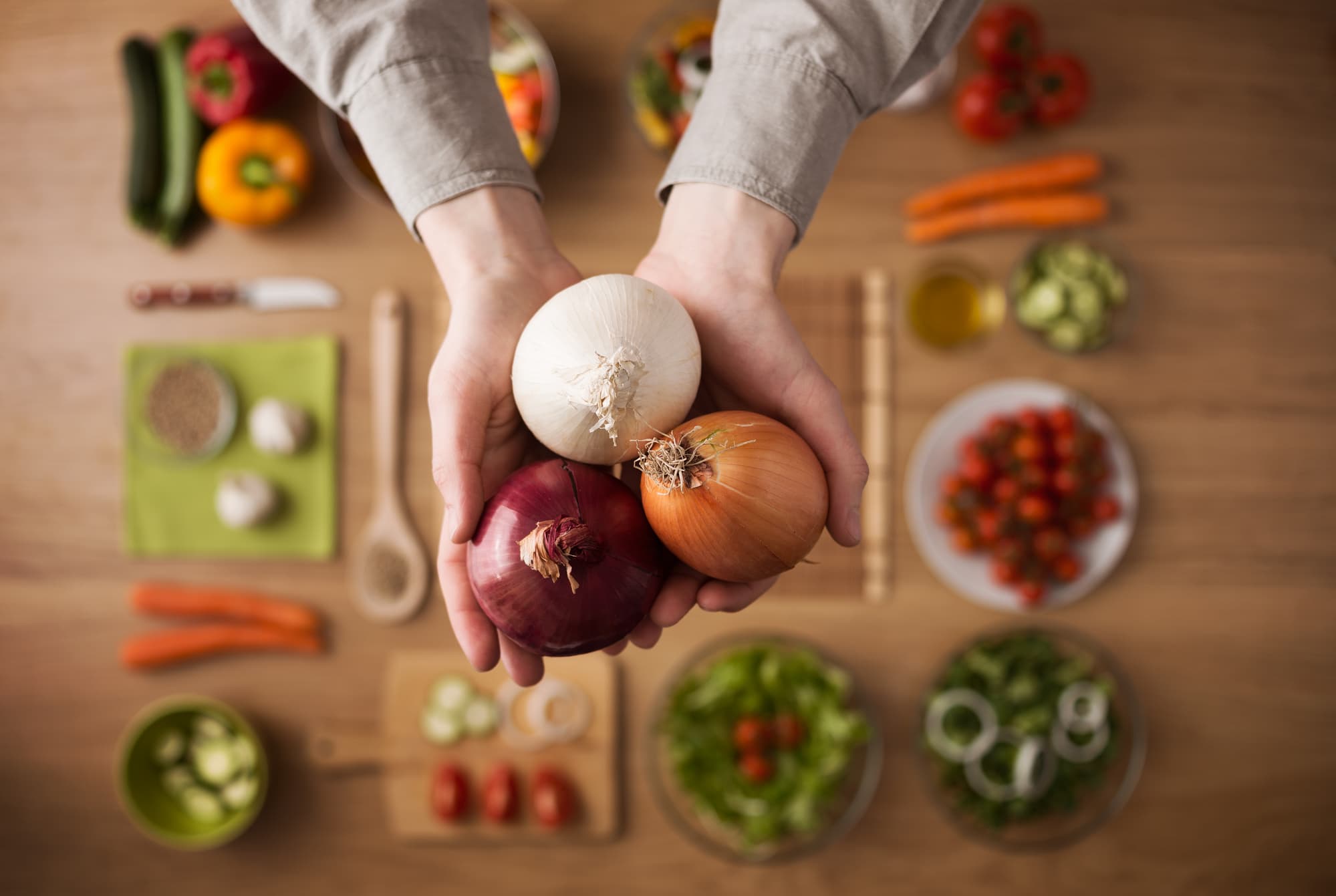 A hand is holding three different type of onions. More ingredients are in the background. 