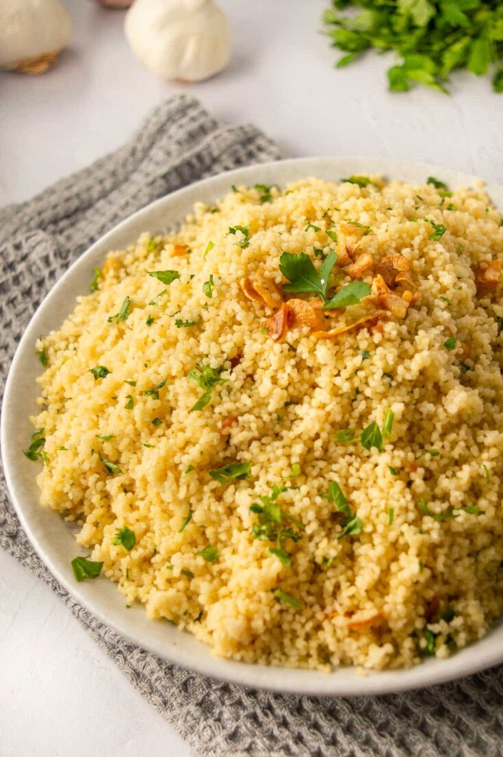 A plate of couscous topped with herbs and brown fried garlic on a grey towel on a white background. 