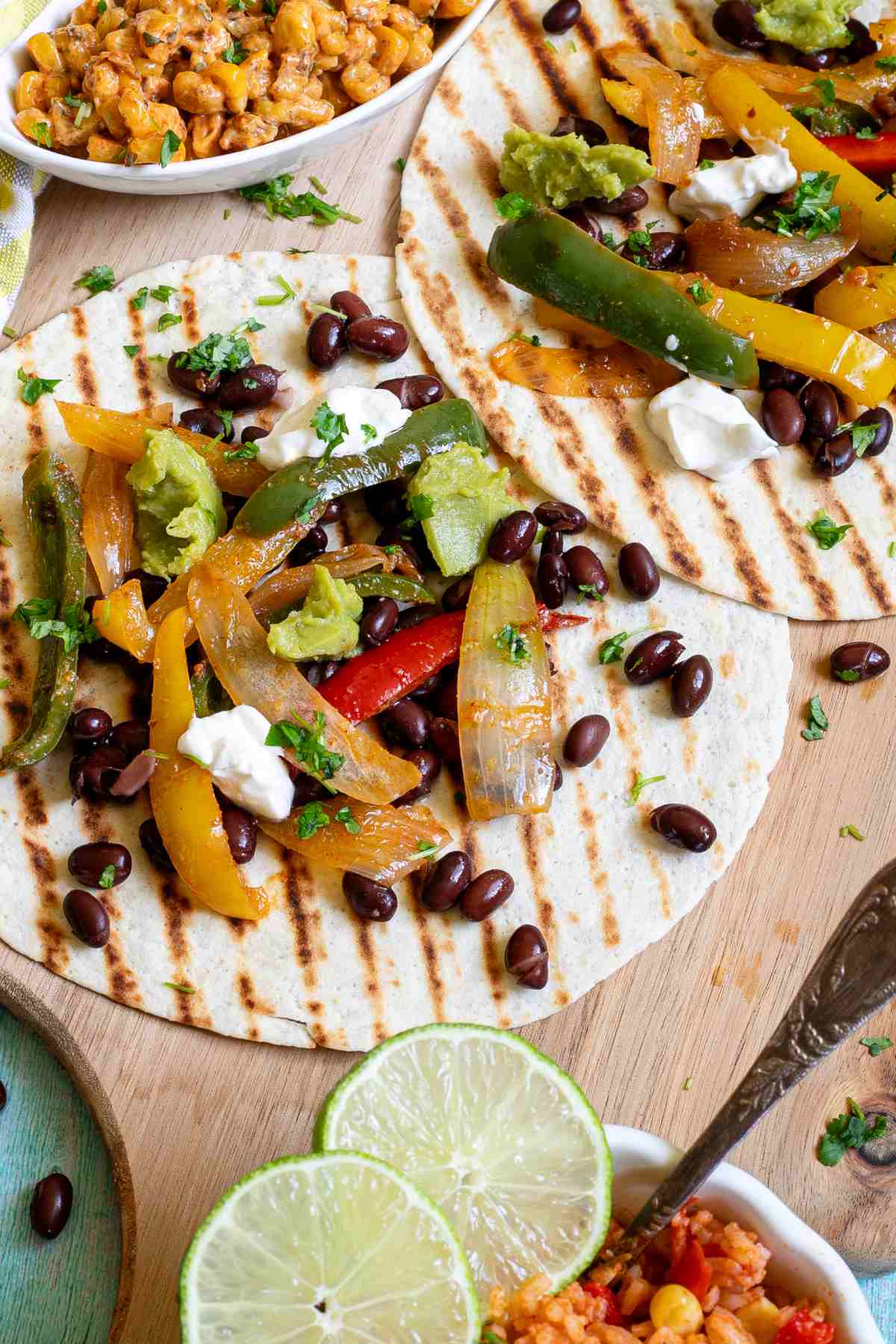 Two tortillas with charred grill lines topped with colorful bell pepper and onion strips as well as black beans, topped with sour cream and guacamole. Corn-based side dishes are in small bowls next to them.