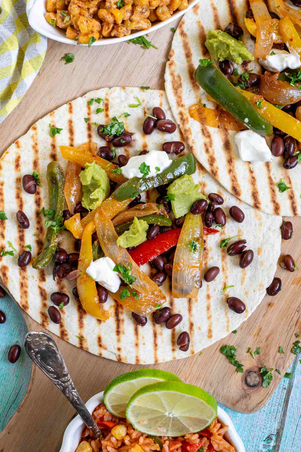 Two tortillas with charred grill lines topped with colorful bell pepper and onion strips as well as black beans, topped with sour cream and guacamole. Corn-based side dishes are in small bowls next to them.