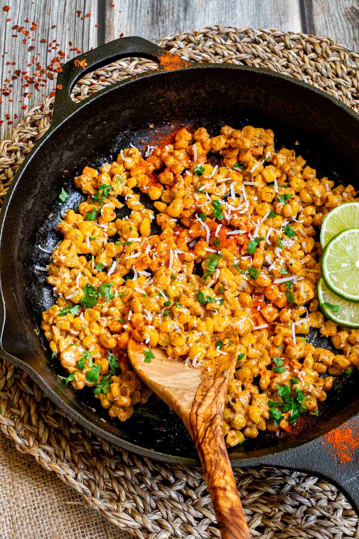 Cast iron skillet with charred yellow corn covered in spices and chopped herbs. A wooden spatula and two lime slices are placed inside.