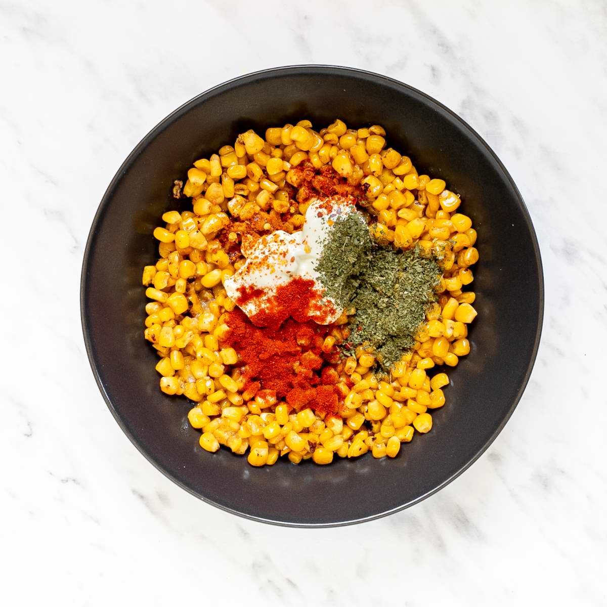 Black bowl full of slightly charred yellow corn kernels and red, green spices, shredded cheese and mayo.