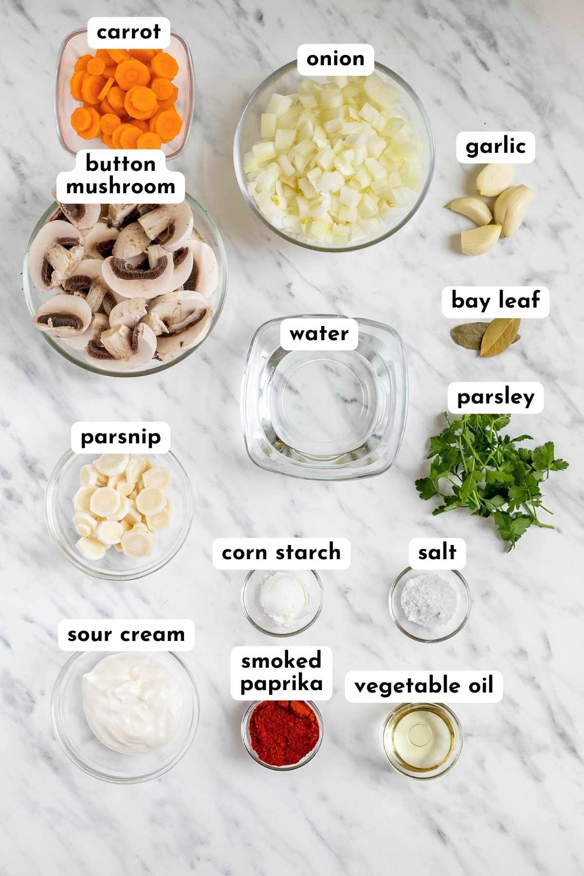 Ingredients of Hungarian mushroom soup in small glass bowls like mushroom, chopped carrots, onion, parsnips, spices, herbs, and other liquid ingredients.