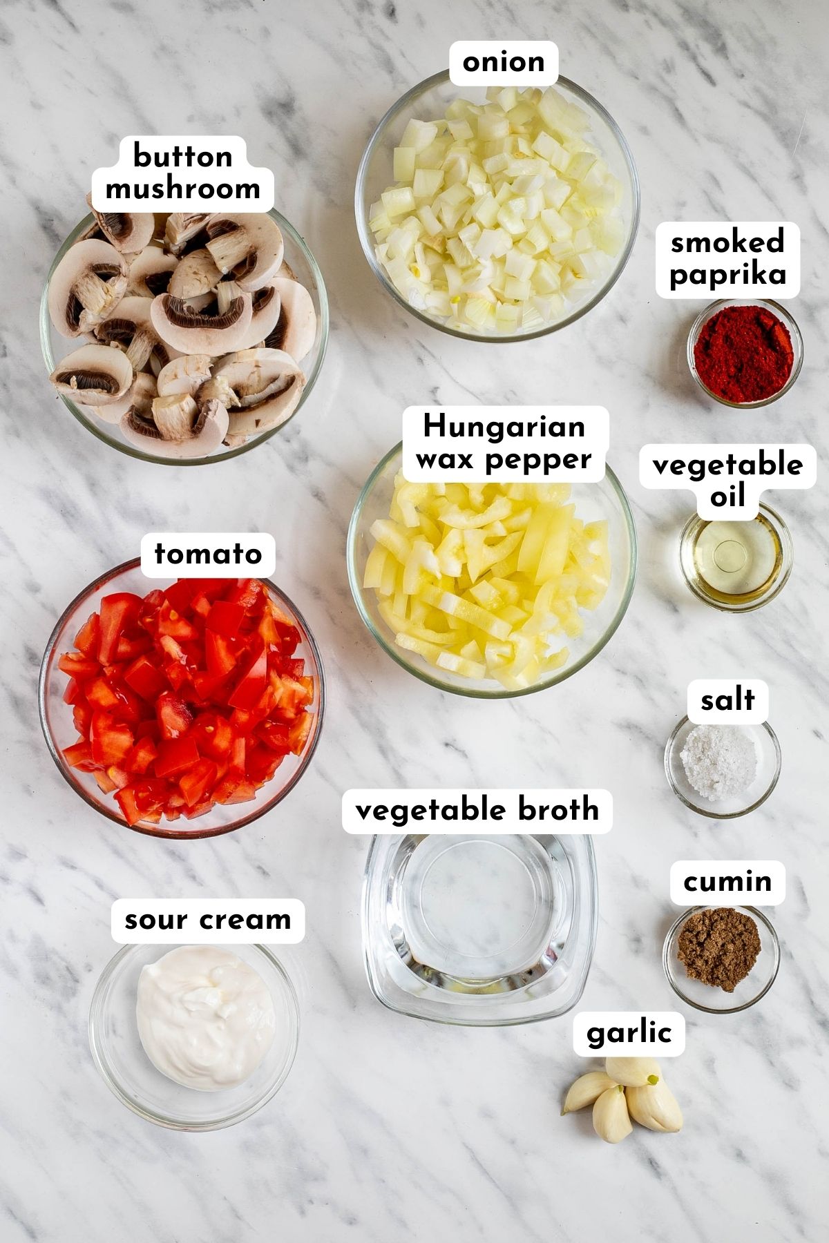 Ingredients of Hungarian goulash in small glass bowls like mushrooms, chopped pepper, tomato, onion, carrot, potatoes, spices, and liquid ingredients.