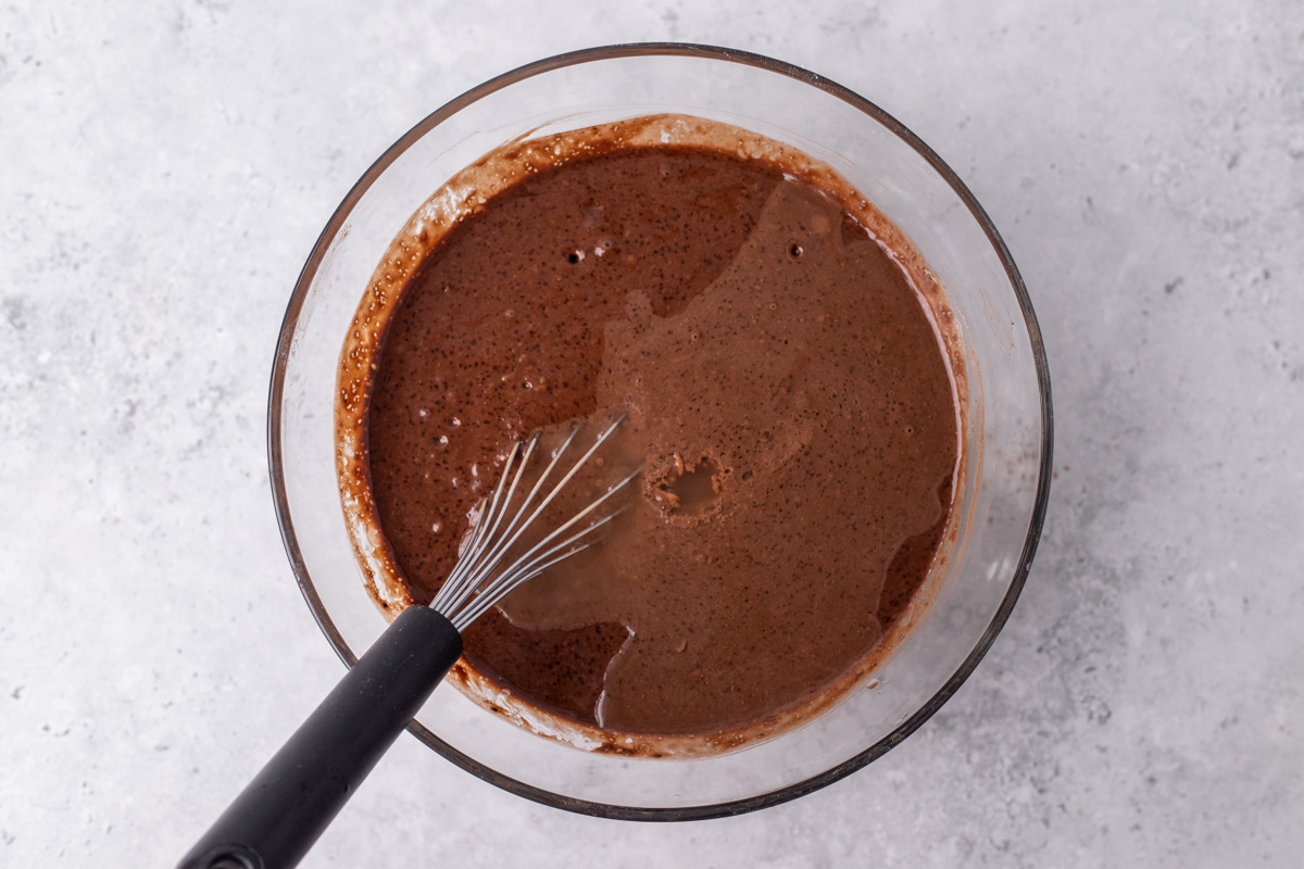 Glass bowl with dark brown cake batter. There is a whisk in it.